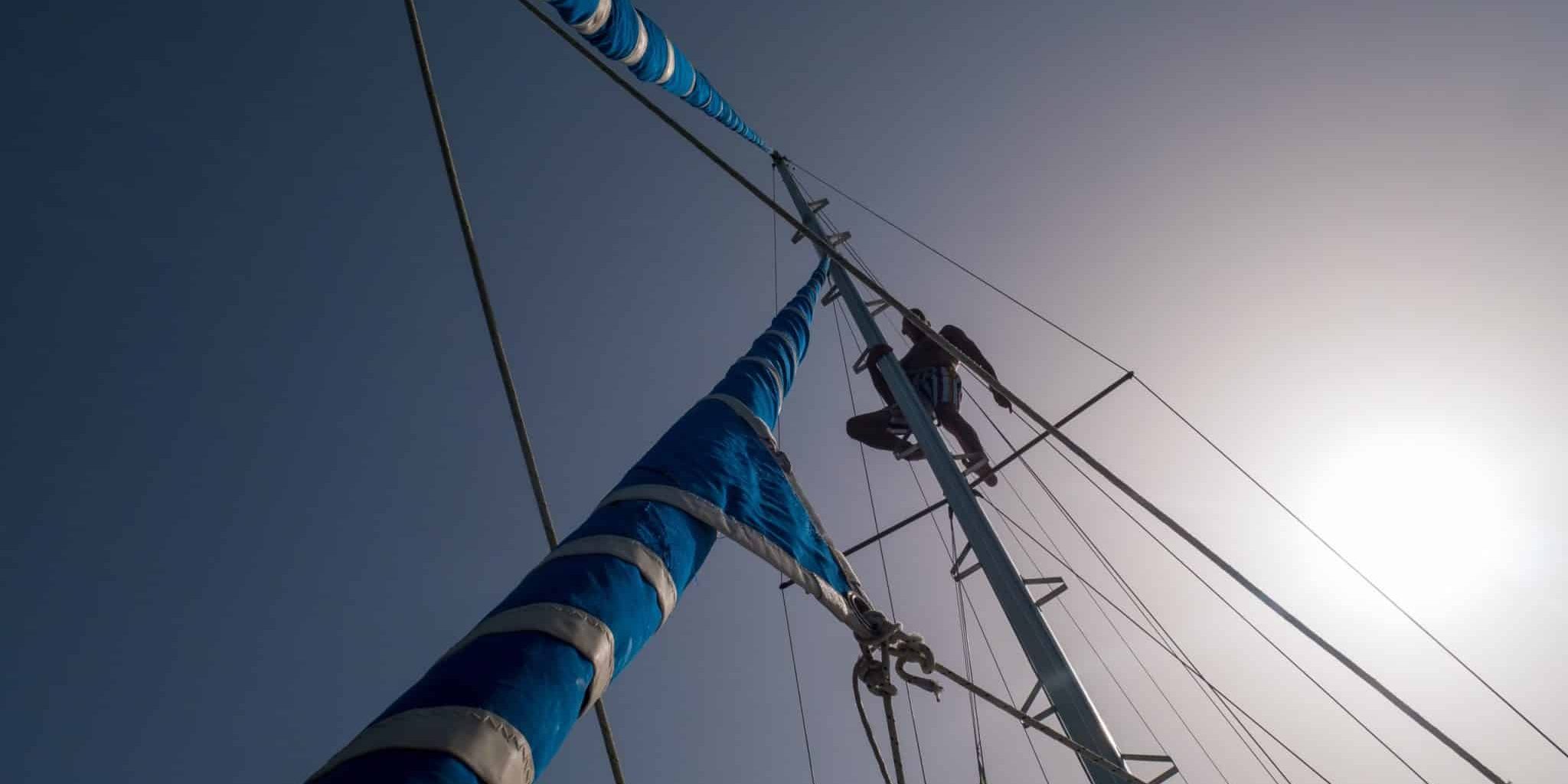 Skipper climbs up on the pole of a sail boat. View from deck up to sky.